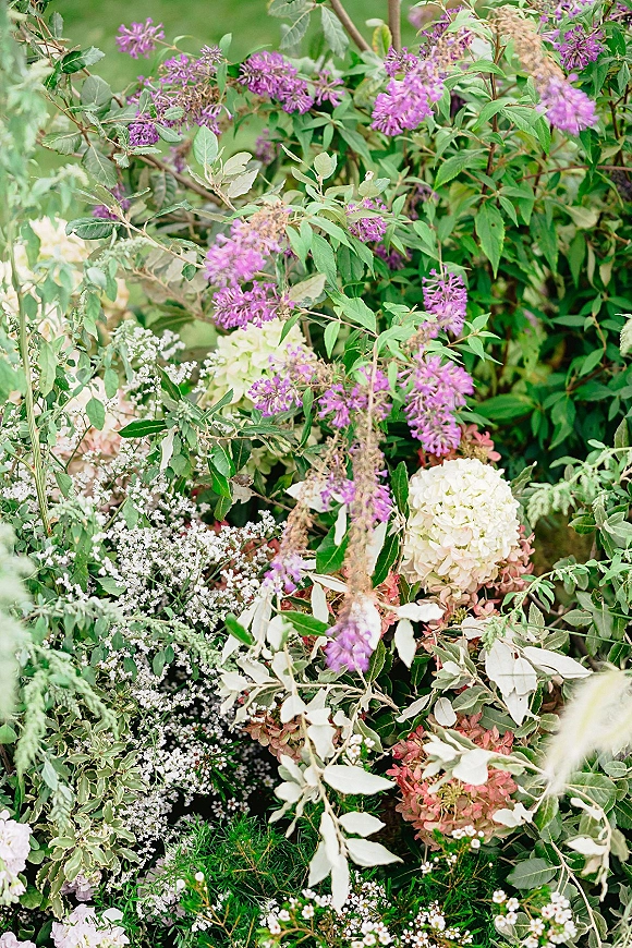Wedding florals with ceremony aisle flowers featuring purple blooms, white and pink hydrangea, and greenery arranged on grass in a garden setting