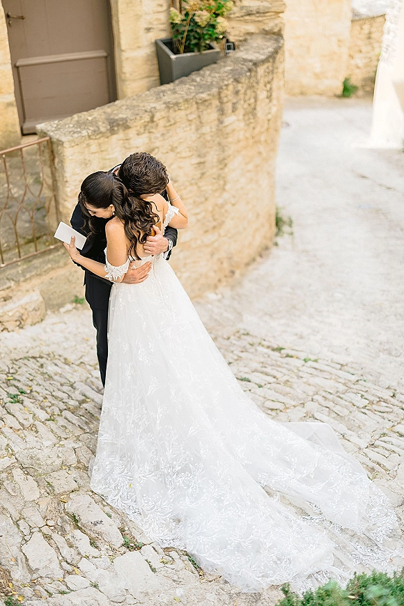First look moment as bride in off-the-shoulder lace gown hugs groom in tux while reading a note on a cobblestone street by stone wall