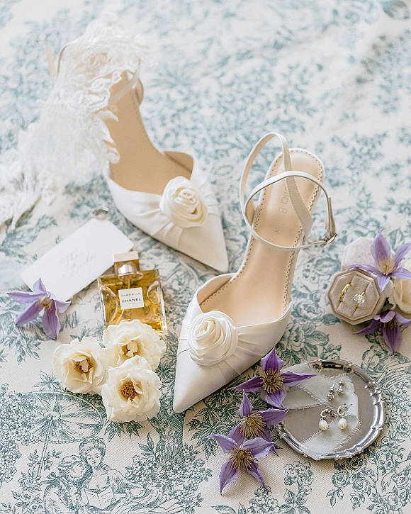 Bridal shoes flatlay with wedding details flat lay, white pointed-toe heels with rosettes beside ring box, perfume, earrings on blue toile fabric