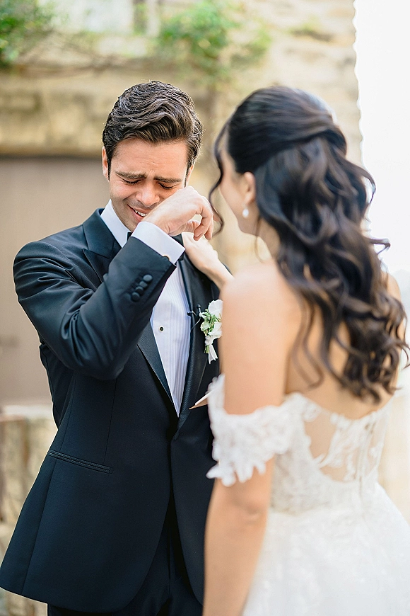 First look moment as bride taps groom’s shoulder; he wipes tears in a black tuxedo beside a stone wall with greenery behind