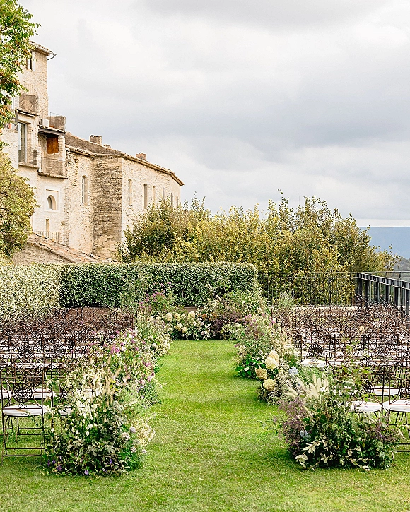 Ceremony aisle decor with greenery garlands and aisle florals beside wrought iron chairs, set on a lawn near a stone building terrace