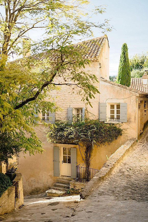 Wedding venue exterior with ivy-covered stone building, blue shutters, and lantern by a cobblestone path beneath trees and cypress sky view