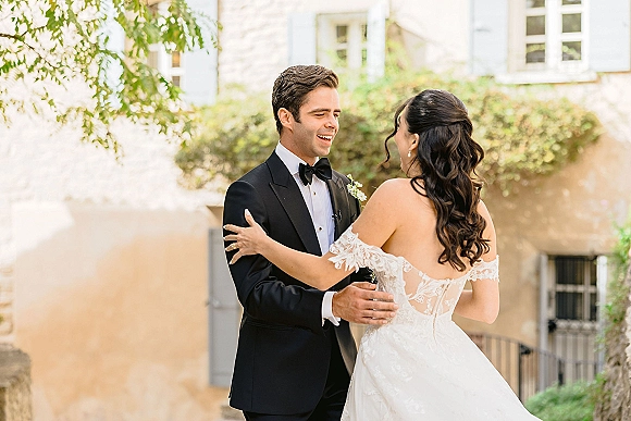 First look moment as bride in a lace-sleeved wedding dress hugs groom in black tuxedo with boutonniere in a stone courtyard with greenery