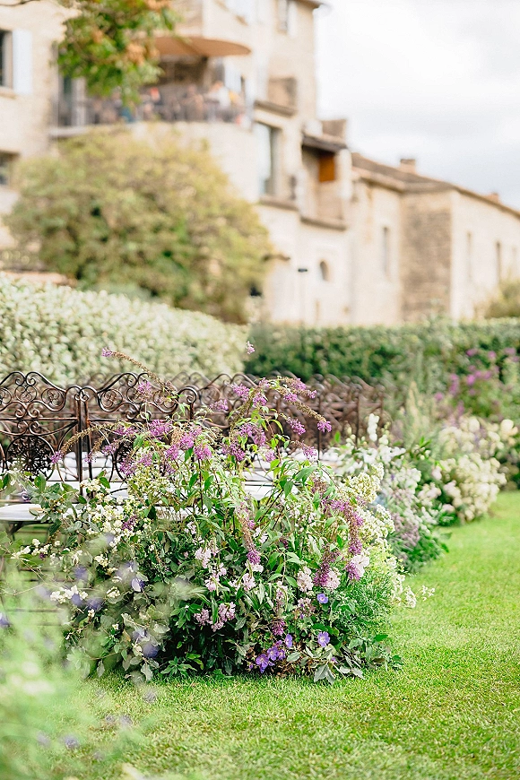 Wedding ceremony seating with wrought iron benches and white floral accents lining a lawn aisle before a stone building with balcony