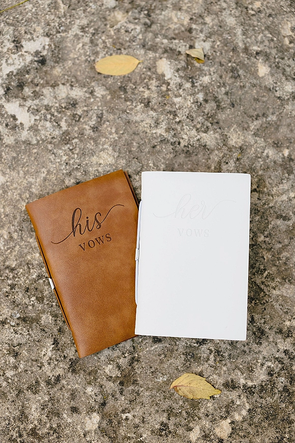 Vow books set with a tan leather vow book and white vow book, metal pen clip, resting on stone ground with fallen leaves