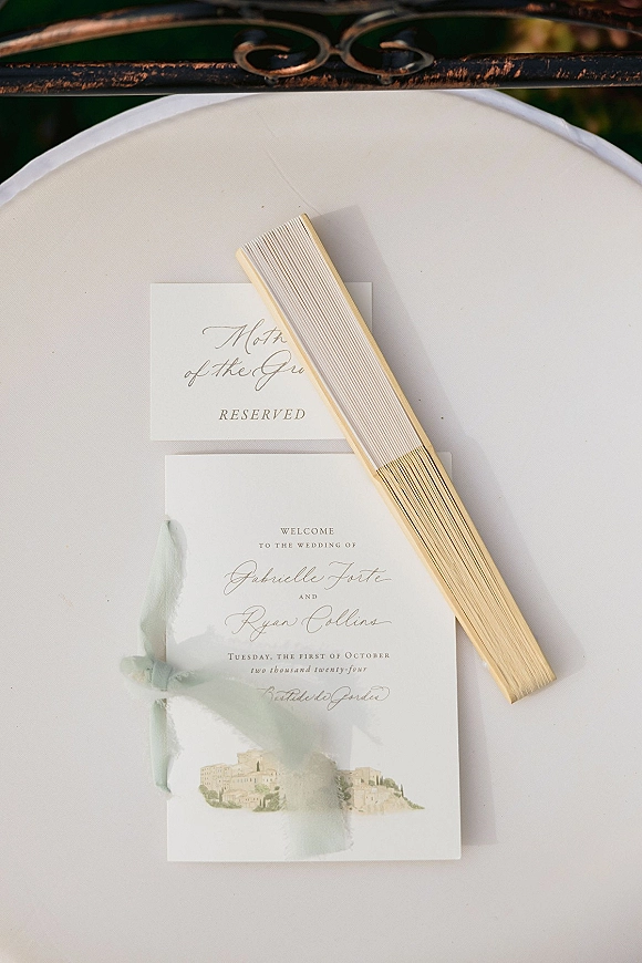 Wedding invitation suite with calligraphy wedding invitations, vellum overlay and silk ribbon, plus reserved seat card and wooden fan on a chair cushion