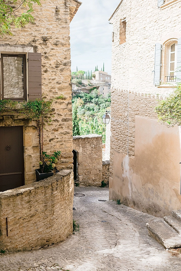 Stone alleyway with old stone village street charm, featuring potted plants, climbing vines, and shutters along sunlit cobblestones