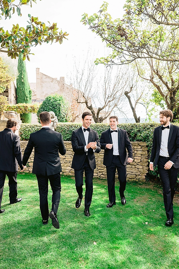 Groomsmen portrait of men in black tuxedos and bow ties walking and laughing on a garden lawn by a stone wall and villa building
