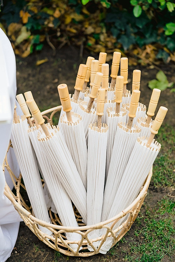 Wedding parasols and white paper parasols with wooden handles in a wicker basket on a garden lawn near a white drape