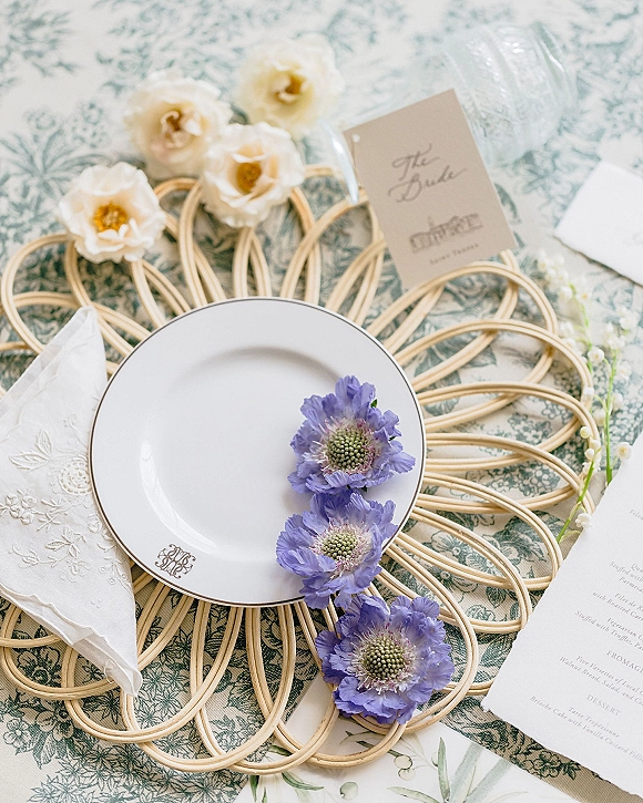 Reception tablescape wedding place setting with rattan placemat, monogram plate, embroidered napkin, menu card, and purple flowers on patterned cloth