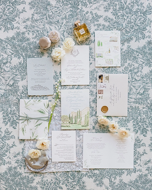 Wedding invitation suite flat lay with vellum wrap and wax seal, calligraphy cards, vintage postage, rings, and white flowers on toile fabric