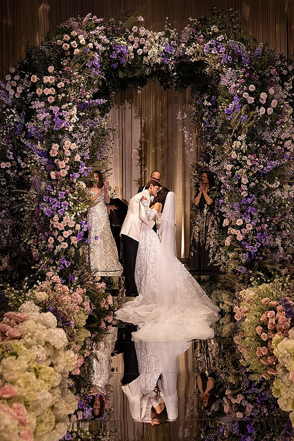 Wedding kiss beneath a floral arch of pink roses and purple blooms, bride in lace gown and veil, groom in white jacket on mirrored aisle indoors