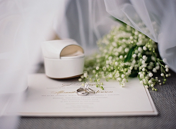 Wedding rings and engagement ring set on a script invitation card beside lily of the valley on soft fabric in gentle light