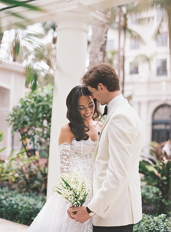 Couple portrait of bride and groom touching foreheads, her off-shoulder lace dress and lily of the valley bouquet in a columned courtyard