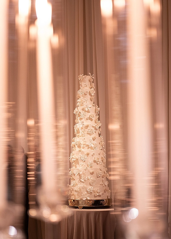 Wedding cake with white fondant and sugar flowers on a pedestal table, set against draped fabric with soft candlelit bokeh indoors