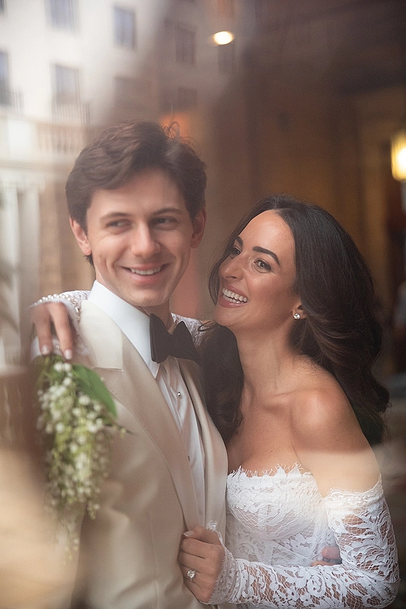 Couple portrait of bride hugging groom in a white tuxedo jacket and black bow tie on an outdoor walkway with warm city lights behind