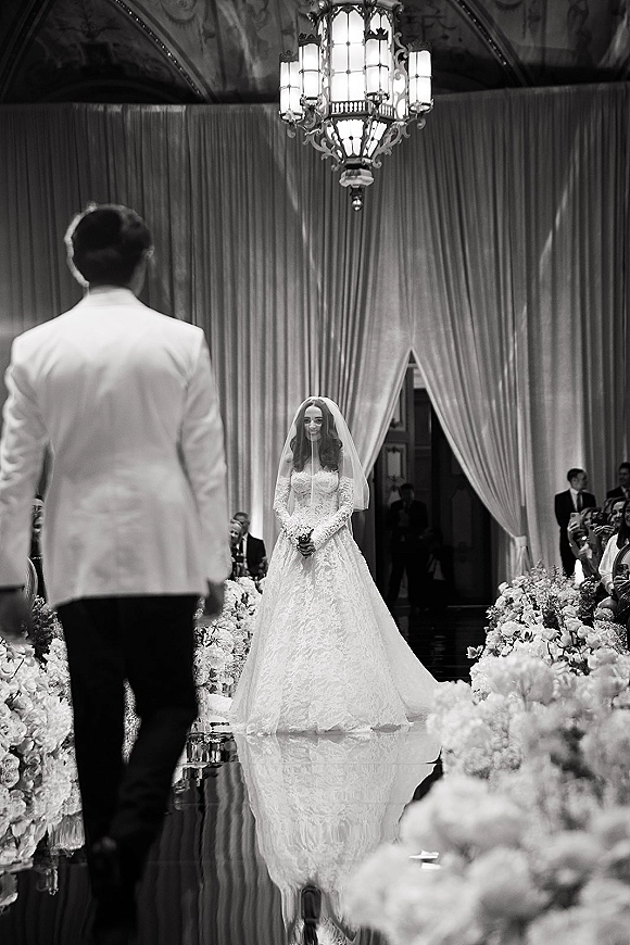 Processional moment as bride walking down aisle in lace gown with long veil, bouquet in hand, toward groom under chandelier in ballroom