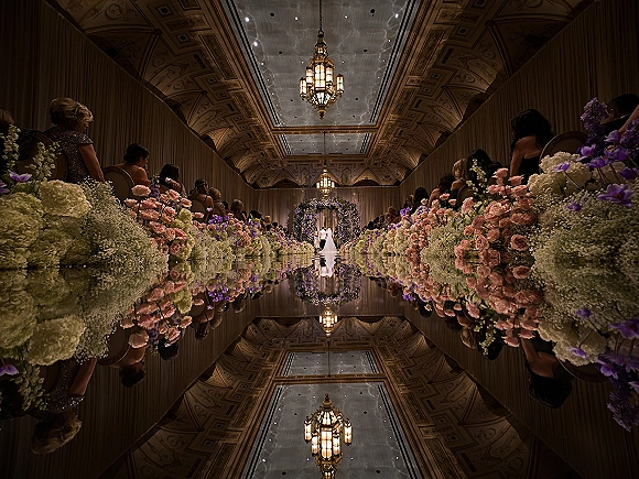 Ceremony aisle design with a mirrored wedding aisle lined in blush roses, hydrangeas and purple blooms beneath chandeliers in an ornate ballroom