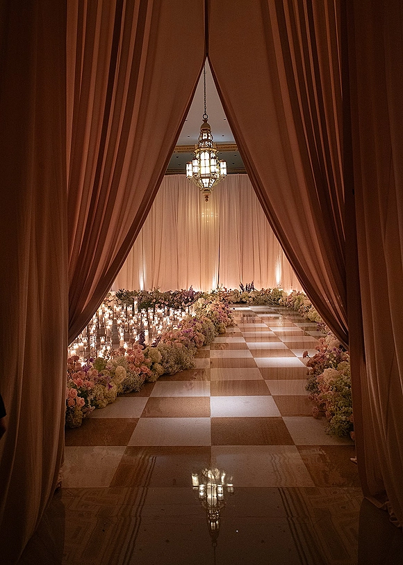 Ceremony aisle decor with a candlelit wedding aisle, low pastel florals and glass votives on a checkered marble ballroom floor beneath chandeliers
