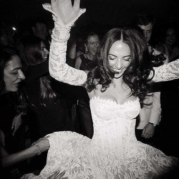 Bride dancing on the wedding reception dance floor with hands raised, wearing a strapless corset bodice as guests cheer in dark lighting