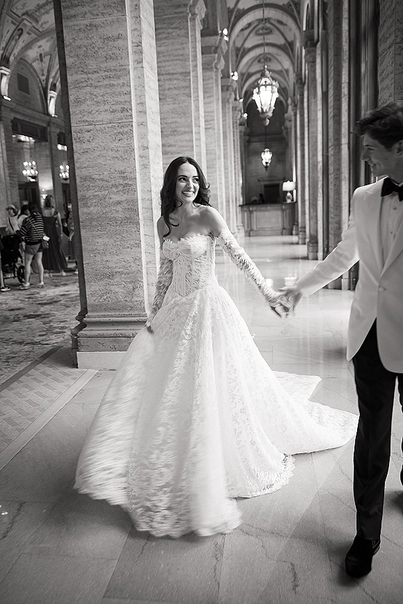 Couple portrait of bride and groom holding hands, walking through an arched hallway with stone columns and chandeliers, her lace dress train flowing