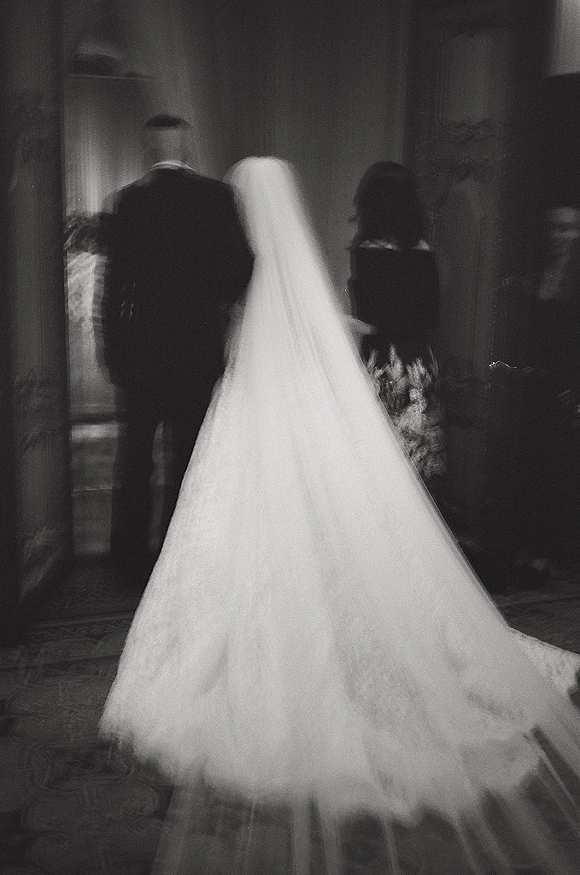 Bride walking down aisle in a wedding processional moment, cathedral veil and long train trailing behind in a stone-walled church interior