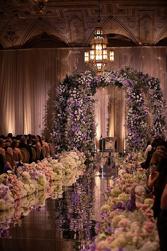 Ceremony aisle design with mirrored wedding aisle, lush floral arch of hydrangeas and roses beneath chandelier, ornate ballroom murals and guests