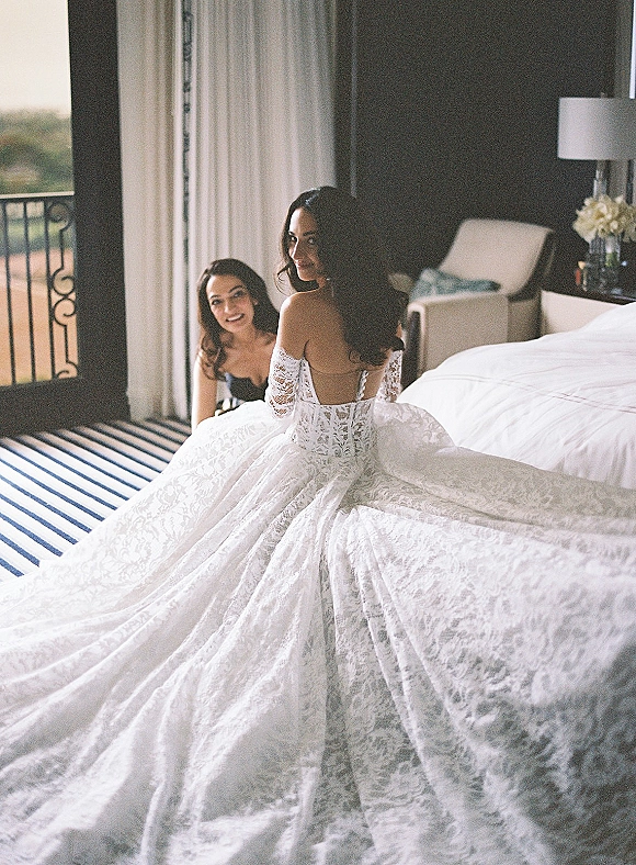 Bride getting ready in a lace wedding dress, sitting on the bed with long train spread behind, softly lit by hotel room window light