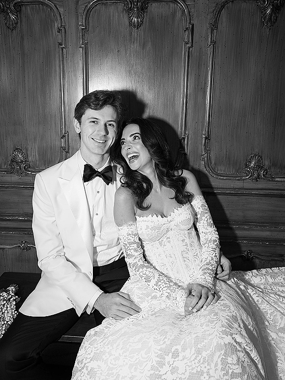 Couple portrait in a black and white wedding portrait style, bride in lace long sleeves laughing beside groom in white tuxedo on bench