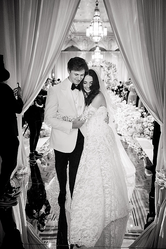 Couple portrait in a black and white wedding portrait, bride and groom embrace on a ballroom aisle beneath chandeliers and draped curtains
