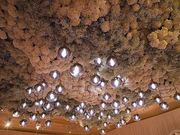 Floral ceiling installation with hanging flower ceiling of hydrangeas, baby's breath, and greenery, accented by glass globe lights and brass rods indoors