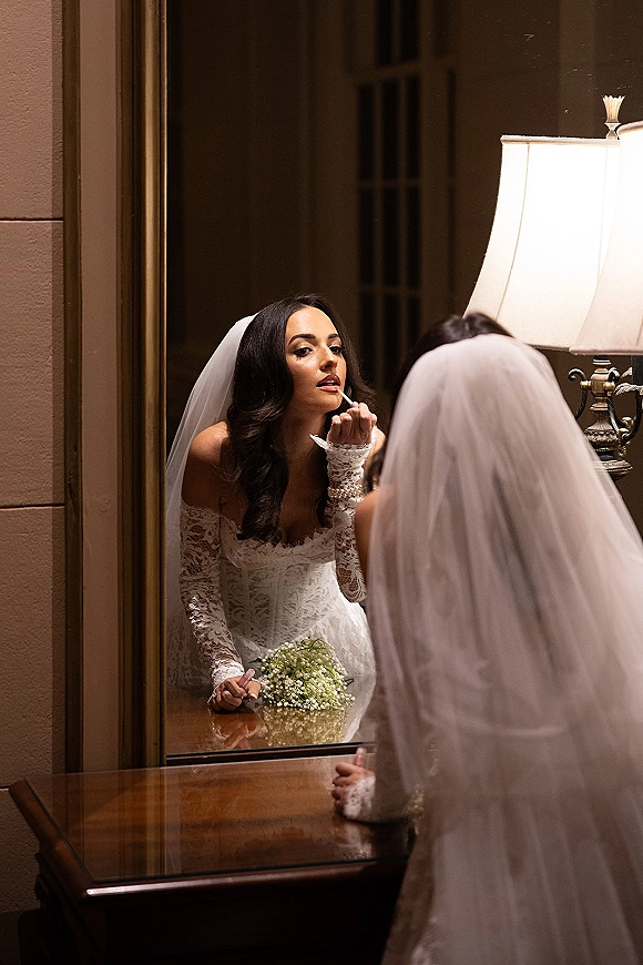 Bridal makeup as bride applying lipstick in mirror, wearing lace off-the-shoulder gown and veil at vanity with lamp and baby's breath bouquet