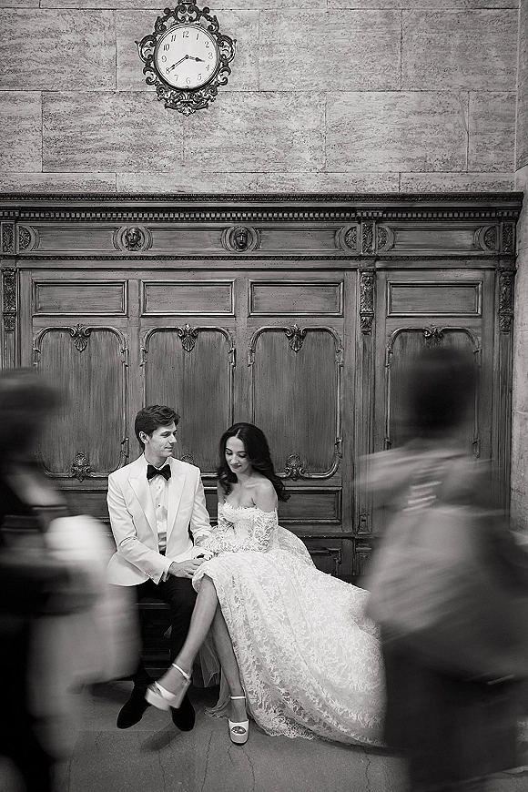 Couple portrait in a black and white wedding portrait, bride in off-shoulder lace gown and veil holding groom’s hand on a bench by a wall clock