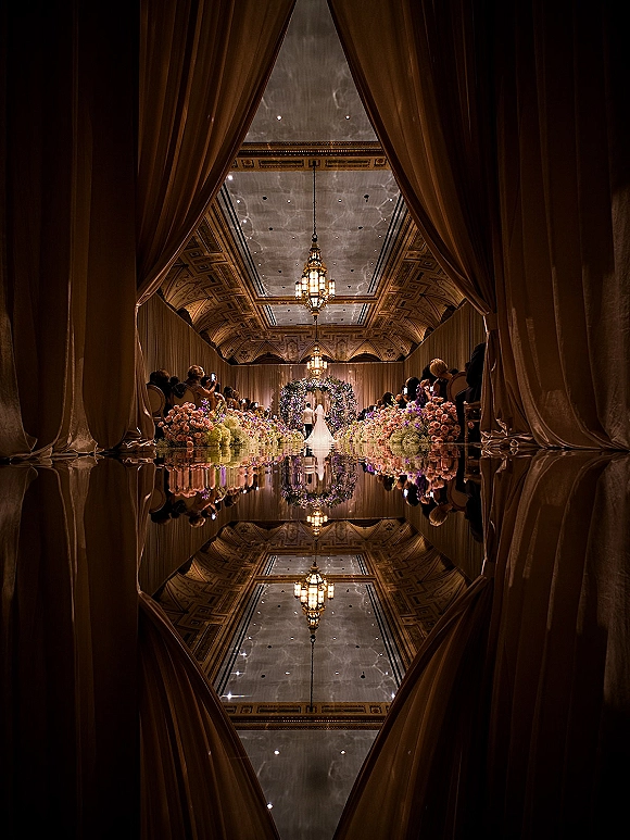 Ceremony aisle design with a mirrored wedding aisle, lined with candles and lush florals beneath chandeliers in an ornate ballroom with guests seated
