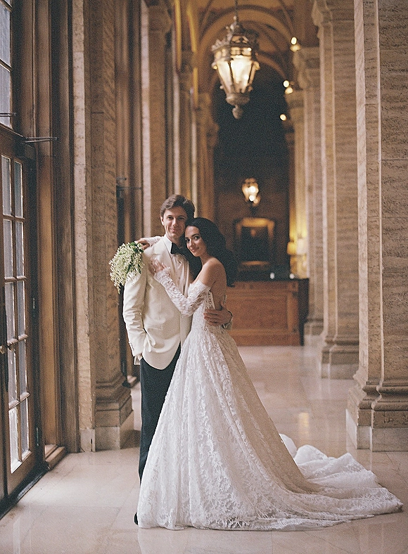 Couple portrait of bride and groom embrace in a stone arched hallway, her lace gown and long train flowing as she holds a bouquet