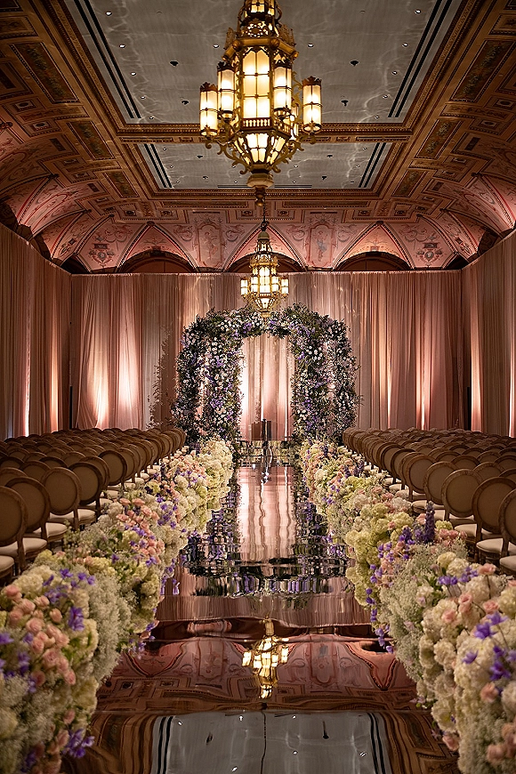 Ceremony aisle design with a mirrored wedding aisle, flower-lined runner, and floral arch under chandeliers in an ornate ballroom ceiling setting