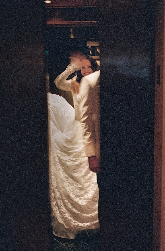 Bride portrait in a long sleeve lace wedding dress with veil, smiling in a dim indoor hallway framed by a doorway and ceiling lights