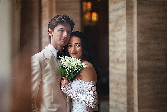 Couple portrait of bride and groom portrait under a stone archway with warm lights, bride holding a baby's breath bouquet in lace gown