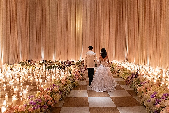 Ceremony aisle decor with candlelit wedding aisle lined by glass hurricane vases and pastel florals as bride and groom walk in a ballroom