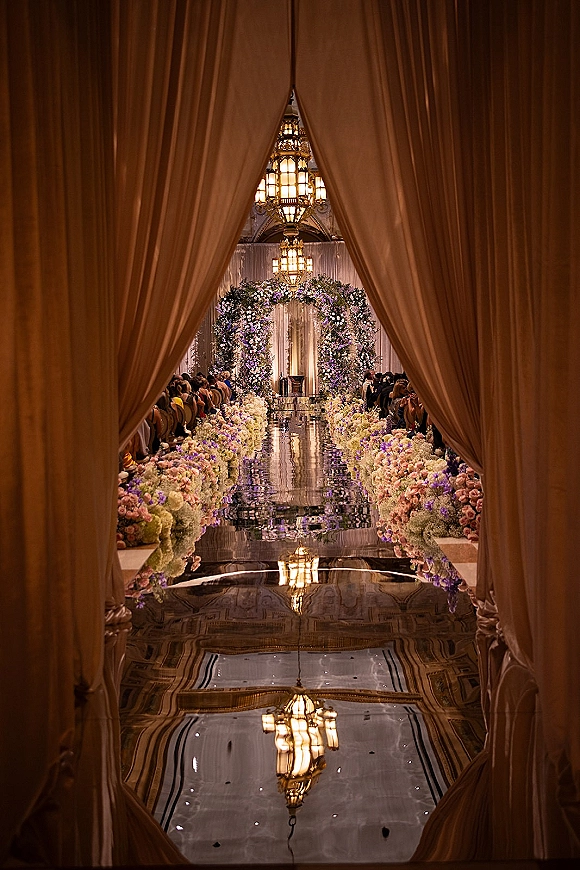 Ceremony aisle design with mirrored wedding aisle runner, lined with floral arrangements and chandeliers in a draped ballroom setting
