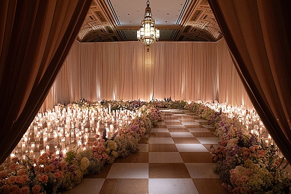 Ceremony aisle decor with a candlelit wedding aisle, roses and hydrangeas beside glass hurricanes, leading to draped ballroom entrance under chandelier