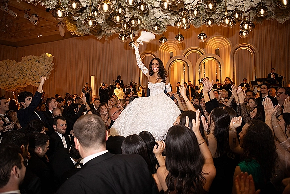 Wedding reception dance as bride in strapless ball gown and lace gloves is lifted on a chair under globe lights in a cheering ballroom crowd