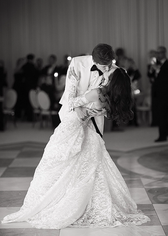 First dance kiss as groom dips bride in long-sleeve lace wedding dress on ballroom dance floor with seated guests watching