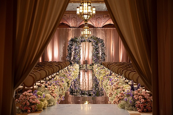Ceremony aisle decor with wedding aisle flowers lining a mirrored runner, leading to a floral arch under chandeliers in an ornate ballroom.