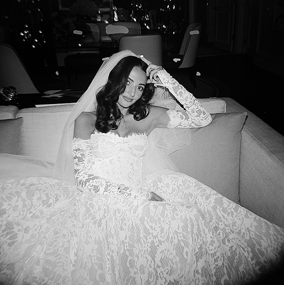 Bridal portrait in black and white of a bride sitting on a sofa in lace off-the-shoulder dress and veil, bokeh lounge lights behind