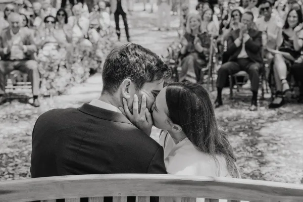 Wedding kiss as the newlyweds share a ceremony kiss moment on a bench, guests applauding along a tree-lined aisle with fallen leaves