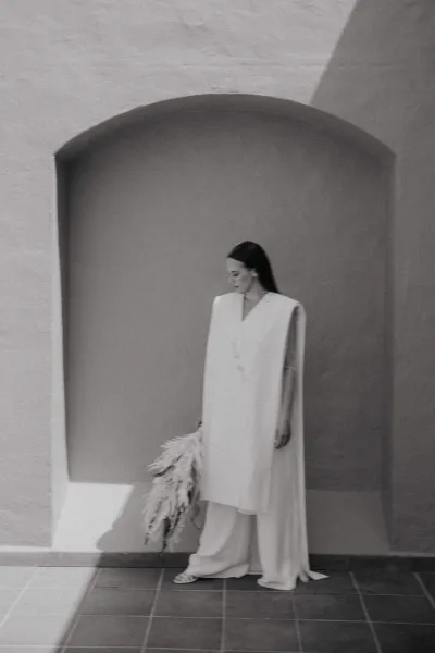 Bridal portrait of a bride in a modern bridal suit with minimalist cape, holding a feather bouquet by an arched niche in hard sunlight shadows