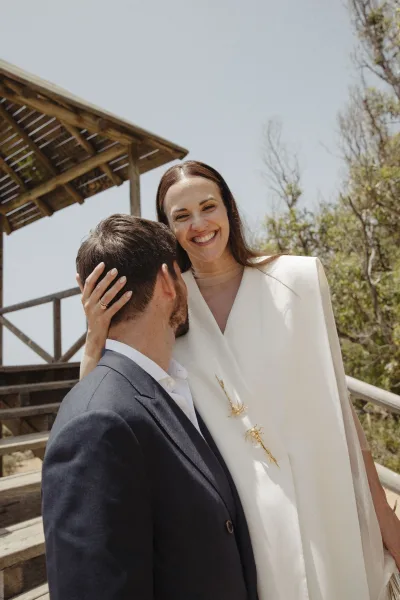 Couple portrait of bride in a white wedding dress and groom in a suit, bride holding his face on wooden stairs by a pergola