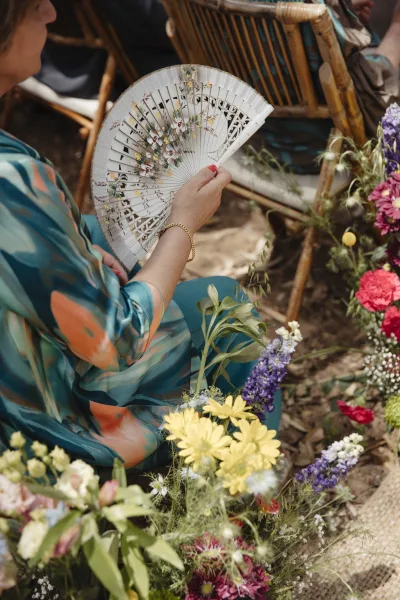 Wedding guest detail with fan, floral dress and gold bracelet beside wicker chair on burlap aisle runner with bright wildflowers accent