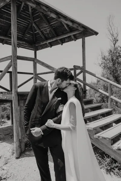 Wedding kiss in a black and white wedding photo, bride in cape sleeve gown and groom in suit by a wooden lookout tower stairs